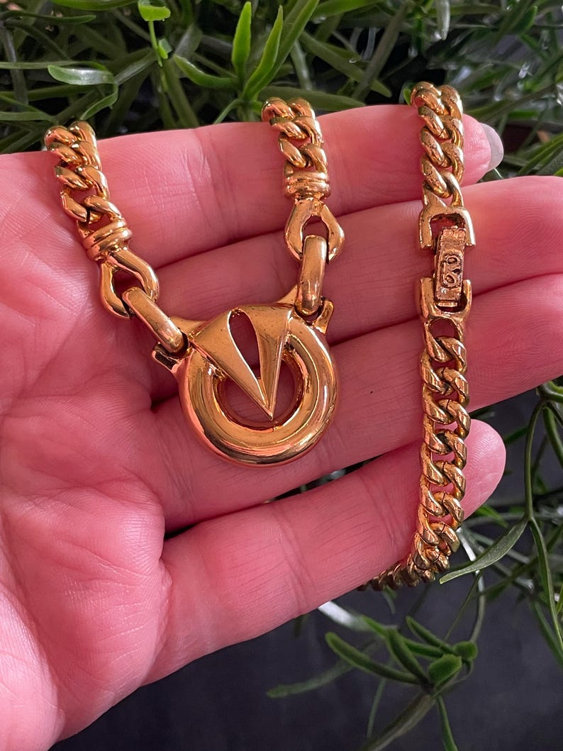 May include: A gold-toned necklace and bracelet set. The necklace features a large circular pendant with a V-shaped design. The chain links are thick and rectangular. The bracelet has a matching chain and clasp. The jewelry is displayed against a green background.