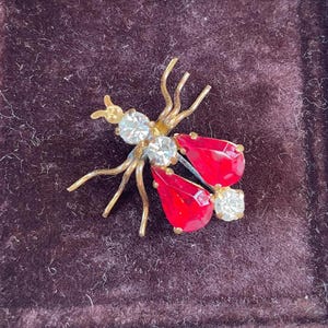 May include: A vintage insect brooch with a gold-tone metal body. The brooch features clear, sparkling rhinestones and two large, teardrop-shaped red stones. The wings and body are detailed with delicate lines.