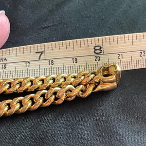 May include: Close-up of a gold-colored chain with a curb link design. The chain is next to a wooden ruler, showing measurements in inches. The chain's clasp is visible, and the background is a dark fabric.
