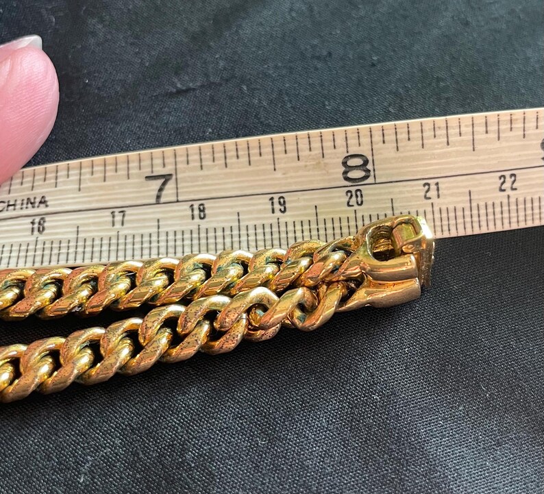 May include: Close-up of a gold-colored chain with a curb link design. The chain is next to a wooden ruler, showing measurements in inches. The chain's clasp is visible, and the background is a dark fabric.