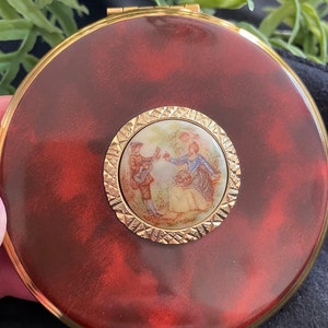 May include: A round, red compact with a gold rim and a gold-rimmed, white porcelain medallion featuring a painted scene of two people in a garden.
