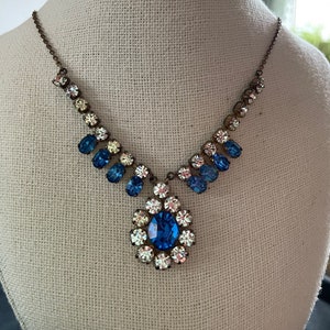 May include: A vintage-style necklace with a large blue rhinestone pendant surrounded by smaller clear rhinestones. The necklace is made of a silver-toned metal chain and features a cluster of smaller blue rhinestones on each side of the pendant.