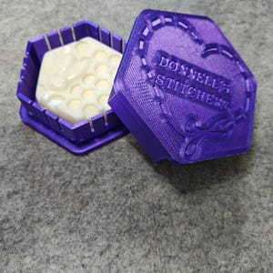 May include: A purple, hexagonal container with a lid, holding a white, honeycomb-shaped item. The lid is open, revealing the contents. The lid has the text "DONNELL'S STITCHERY" and decorative stitching.