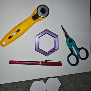May include: A yellow OLFA rotary cutter, teal scissors, a red pen, a multi-colored hexagon-shaped object, and two white hexagon-shaped paper cutouts are arranged on a white surface.