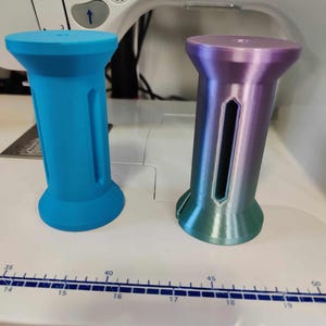 May include: Two cylindrical objects with a flared base and top. One is solid turquoise, the other is a gradient of purple, blue, and green. Both have a central cutout. They are placed on a white surface with measurement markings.