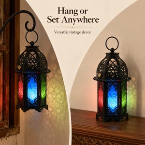 May include: Two black metal lanterns with intricate cut-out patterns and colored glass panels in red, blue, and green. One lantern hangs from a curved metal hook, while the other sits on a flat surface. Text reads: Hang or Set Anywhere.