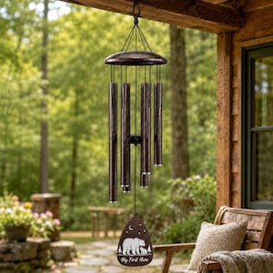 May include: A bronze-colored metal wind chime featuring five cylindrical tubes of varying lengths suspended from a circular top. A teardrop-shaped wooden wind catcher hangs at the bottom, displaying a white graphic of two bears and the text My First Hero.