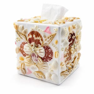 May include: A square white tissue box cover decorated with an assortment of natural seashells, including brown fan shells, white spiral shells, and a central starfish. The design is accented with small white pearls and pink shell fragments.