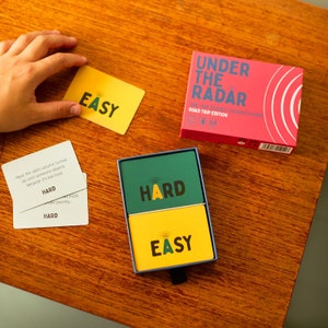 May include: A card game set on a wooden surface, featuring a red box with the text "UNDER THE RADAR". The game includes cards labelled "EASY" and "HARD". Some cards are in a blue box, others are loose.