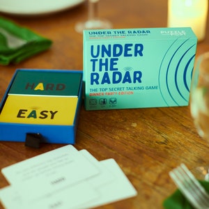 Puede incluir: Una caja turquesa y una caja azul con cartas del juego "Under the Radar: The Top Secret Talking Game Dinner Party Edition" están sobre una mesa de madera. La caja turquesa tiene el título del juego en letras blancas grandes. La caja azul tiene "HARD" y "EASY" en las cartas.