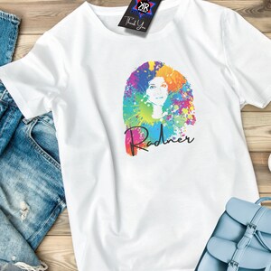 May include: White t-shirt featuring a colorful graphic of a woman's face with vibrant paint splatter hair and the name "Radner" in script. A black tag with the text "Thank You" hangs from the shirt. A light blue purse and denim jeans are also visible.
