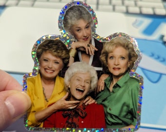 May include: A holographic sticker featuring four women from the Golden Girls television show. The women are wearing colorful outfits, including yellow, red, and green. The sticker has a glittery border.
