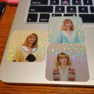 May include: Three holographic stickers featuring a woman in different outfits. One sticker shows her in a yellow sailor-style top with a black bow. Another shows her in a blue top. The third shows her in a white shirt and patterned vest.