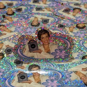 May include: A collection of holographic stickers featuring a woman holding a black video game console. The stickers have a glittery, iridescent finish with a floral and swirl design. The woman is smiling and wearing a cream-colored blazer.