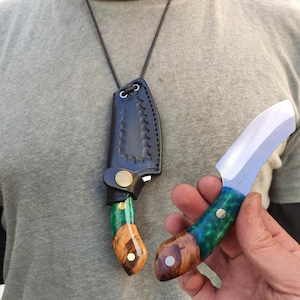 Könnte beinhalten: Ein kleines, handgefertigtes Messer mit gebogener Klinge und einem Griff aus grünem und braunem Holz. Das Messer wird mit einer schwarzen Lederscheide an einer Schnur präsentiert. Der Griff hat einen Messingstift.