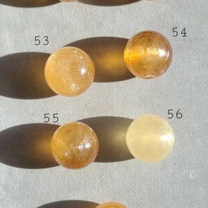 May include: Seven smooth, round, yellow-orange citrine spheres, numbered 51 through 57, arranged in two rows on a light grey surface.