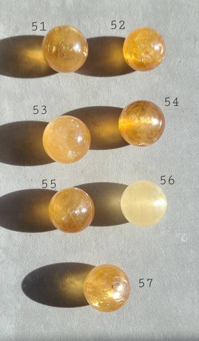 May include: Seven smooth, round, yellow-orange citrine spheres, numbered 51 through 57, arranged in two rows on a light grey surface.