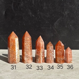Op de afbeelding: Zes zonnesteenpunten in verschillende maten, variërend van ongeveer 5 cm tot 2,5 cm hoog. De punten zijn rood-oranje van kleur met een glinsterende, iriserende afwerking.