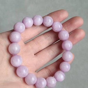Peut inclure: Un bracelet compos&eacute; de perles rondes de couleur lavande, chaque perle mesure environ 11 mm de diam&egrave;tre. Le bracelet est sur une main, et le texte "11mm" est visible sous le bracelet.