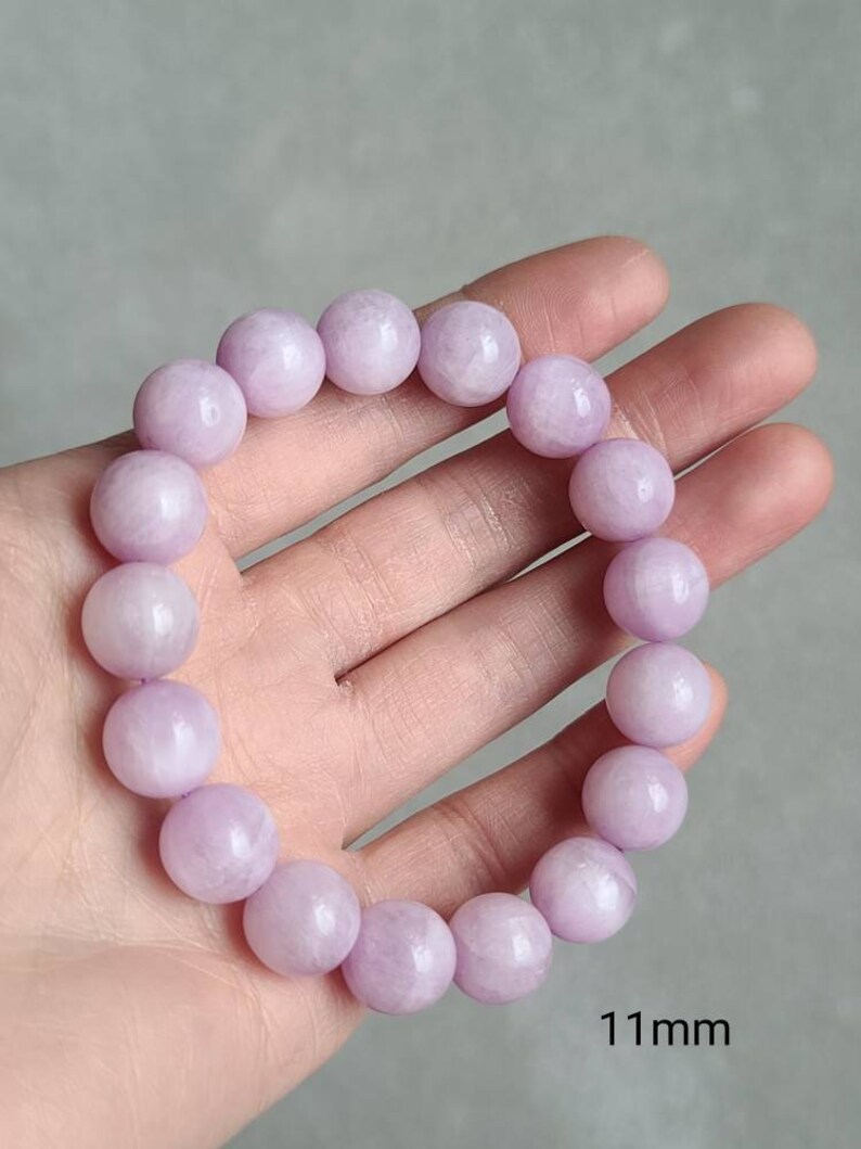 Peut inclure: Un bracelet compos&eacute; de perles rondes de couleur lavande, chaque perle mesure environ 11 mm de diam&egrave;tre. Le bracelet est sur une main, et le texte "11mm" est visible sous le bracelet.