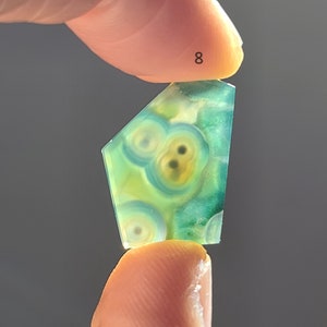 May include: A green and yellow agate cabochon with a unique pattern of circles and swirls. The cabochon is shaped like a teardrop and is held between two fingers. The number 8 is written on the finger holding the top of the cabochon.
