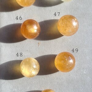 May include: A collection of 7 smooth, round, polished citrine spheres, each with a unique golden hue. The spheres are numbered 44, 45, 46, 47, 48, 49, and 50.