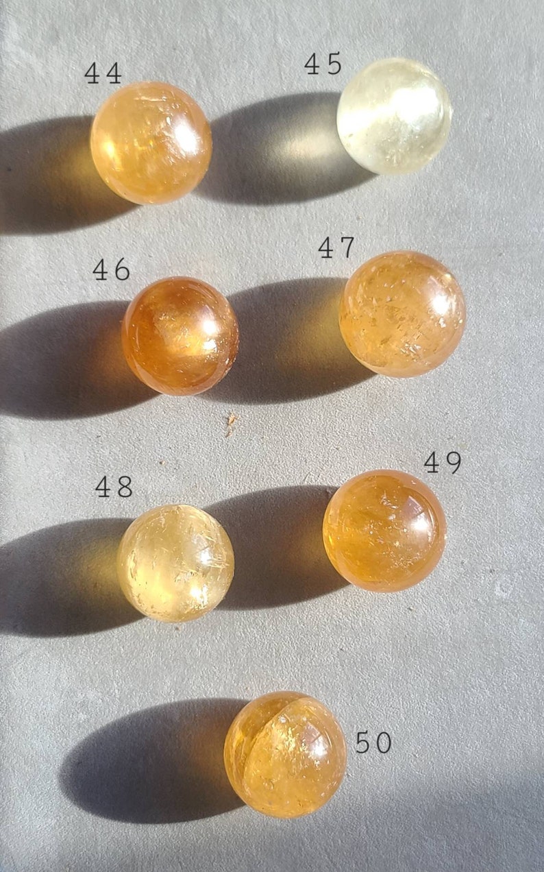 May include: A collection of 7 smooth, round, polished citrine spheres, each with a unique golden hue. The spheres are numbered 44, 45, 46, 47, 48, 49, and 50.