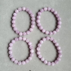 Peut inclure: Quatre bracelets de couleur lavande faits de perles rondes, chaque bracelet mesure 9 mm de diam&egrave;tre. Les bracelets sont dispos&eacute;s en carr&eacute; sur une surface grise.