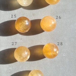 May include: Seven smooth, round, polished citrine spheres, numbered 23 through 29, arranged in rows on a grey surface.