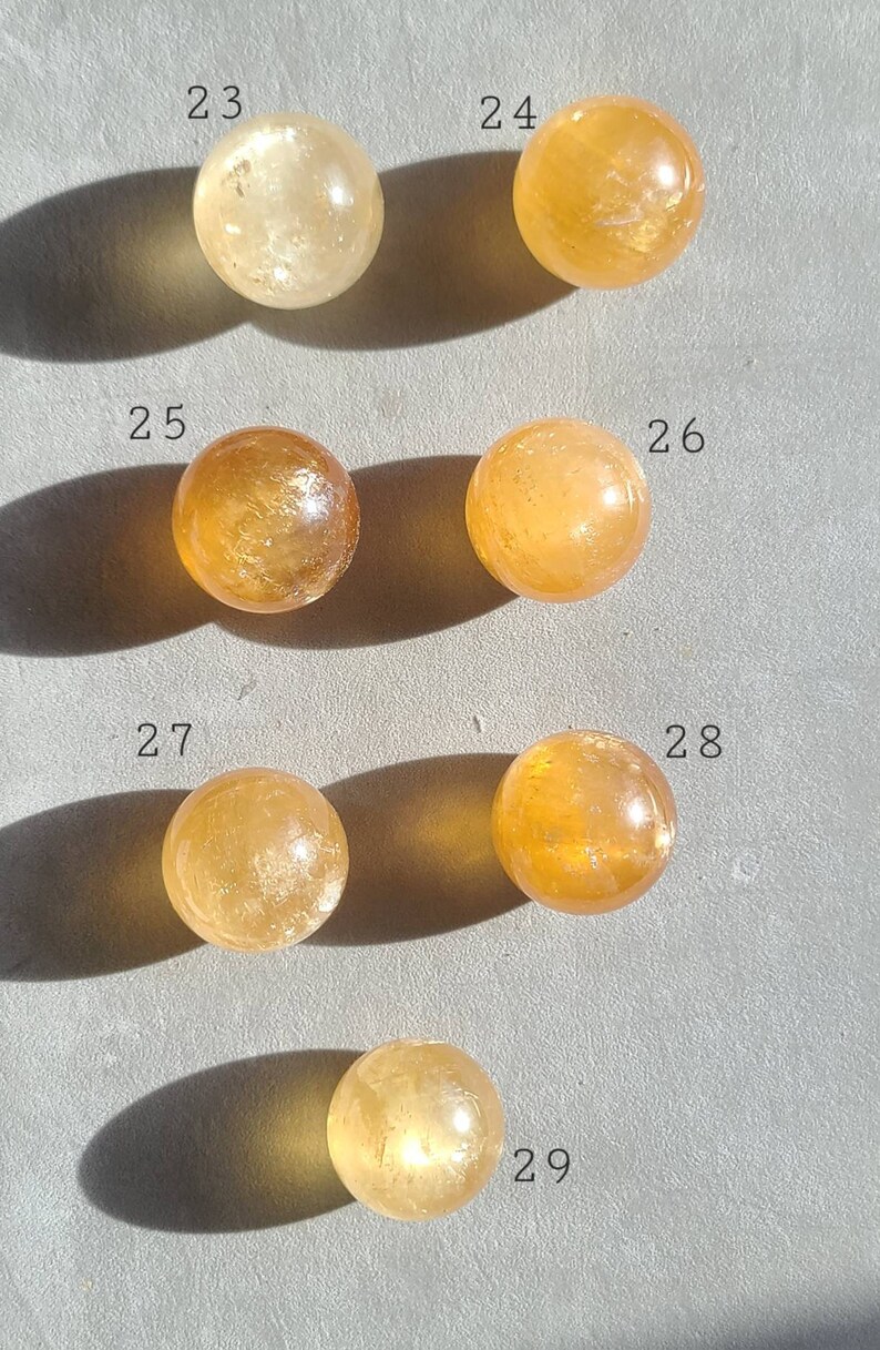 May include: Seven smooth, round, polished citrine spheres, numbered 23 through 29, arranged in rows on a grey surface.