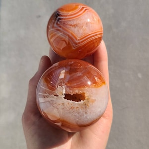 May include: Two Carnelian spheres, one with a natural druzy interior, the other with a smooth, polished surface. Both spheres feature orange and white banding.