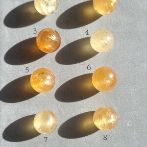 May include: Eight smooth, round, polished citrine spheres, numbered 1 through 8, arranged in two rows of four on a grey surface. The spheres range in colour from light yellow to orange.