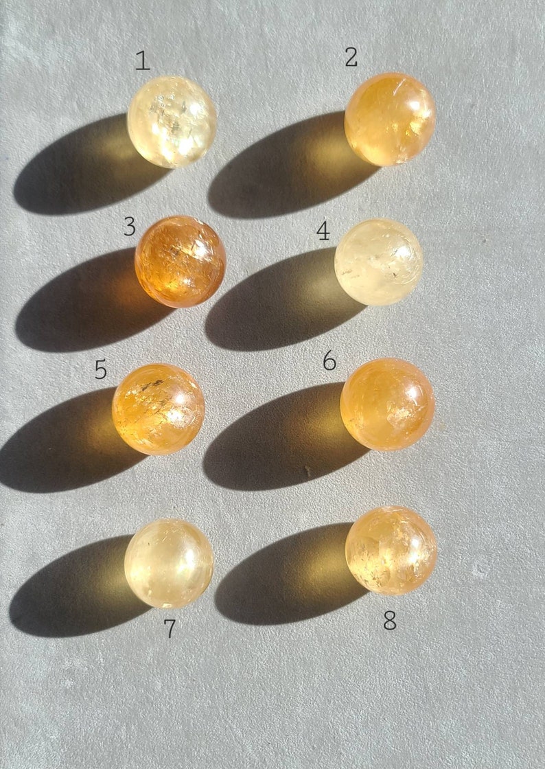 May include: Eight smooth, round, polished citrine spheres, numbered 1 through 8, arranged in two rows of four on a grey surface. The spheres range in colour from light yellow to orange.