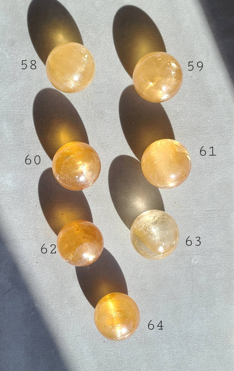 May include: Seven polished, golden yellow, crystal spheres, numbered 58 through 64, arranged in a grid pattern on a grey surface.