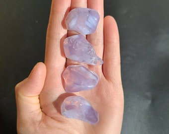 May include: Four polished, light purple gemstones are held in an open hand against a dark background. The stones have a translucent quality, with subtle variations in color and texture. The gemstones are smooth and rounded, with a natural, organic shape.