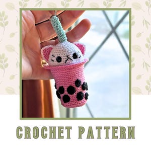 May include: A handmade crochet keychain shaped like a pink boba tea drink with a cat face on top. The keychain has black boba pearls, a white cat face with pink ears, and a green straw. The text "CROCHET PATTERN" is at the bottom.