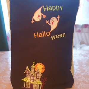 Könnte beinhalten: Schwarze Stofftasche mit grünem und gelbem Besatz. Die Tasche hat ein gesticktes Design mit zwei Geistern und dem Text "Happy Halloween". Der Boden der Tasche hat ein gesticktes Design mit einem Haus und einem Vollmond.