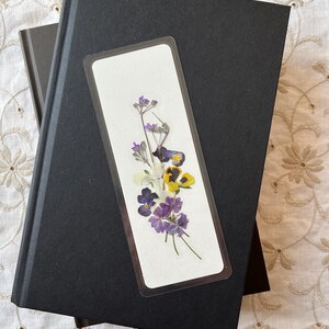 May include: Two black hardcover journals with a floral bookmark. The bookmark features pressed purple, yellow, and white flowers on a white background. The journals are stacked on a white lace tablecloth.