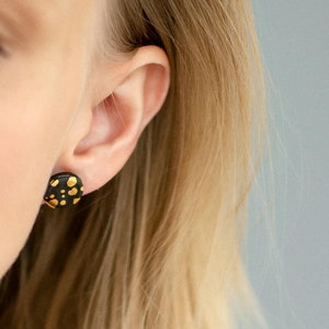 Könnte beinhalten: Ein Paar schwarze Ohrstecker mit goldenen Punkten. Die Ohrstecker sind rund und haben ein gesprenkeltes Muster aus goldenen Punkten auf schwarzem Hintergrund.