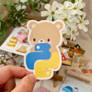 Puede incluir: Una pegatina de un oso marrón con un logotipo de Python azul y amarillo. El oso está sosteniendo el logotipo en sus patas. La pegatina está sobre un fondo blanco.