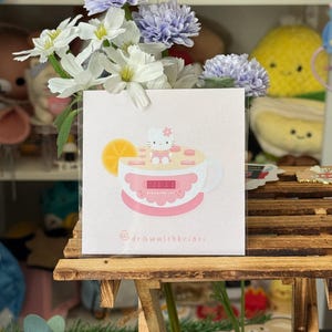 May include: A square print with a cartoon illustration of Hello Kitty in a teacup with a lemon slice. The print has a pink and white colour scheme and the text "@drawwithkristi". The print is displayed with white and purple flowers.