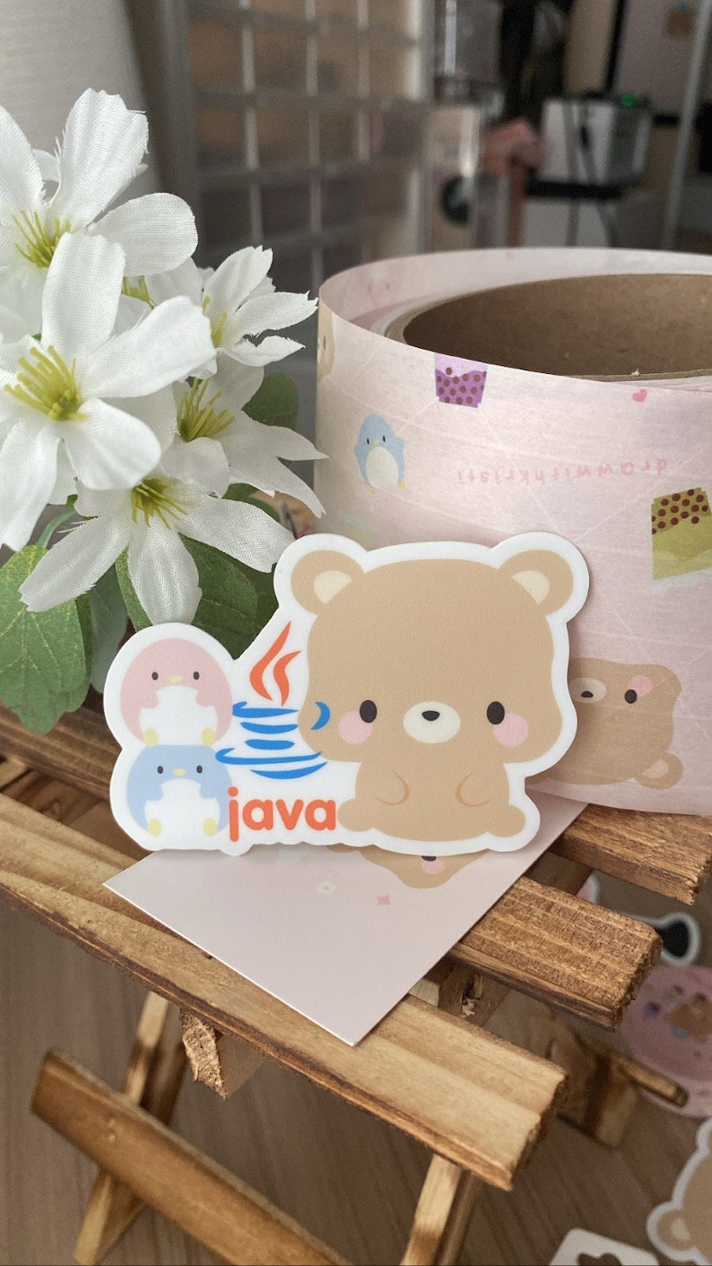 May include: A sticker featuring a cartoon bear holding a Java logo, with two penguin characters. The sticker is light brown with pink cheeks. The background includes a roll of pink patterned paper and white flowers.