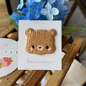 May include: A brown teddy bear patch with a textured, fuzzy appearance. The bear has black eyes, a tan nose, and pink cheeks. The patch is on a white card with the text "@drawwithkristi".