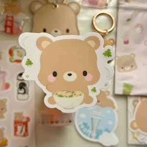 May include: A collection of cute bear-themed stickers and keychains. The central sticker features a brown bear holding a bowl of food, with shamrocks. Other items include a bear keychain, a pink pouch, and stickers with bubble tea and other designs.