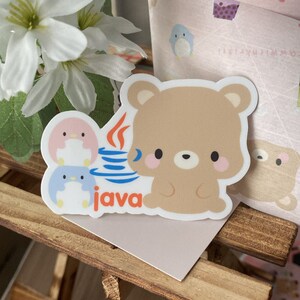 May include: A sticker featuring a cartoon bear and two penguins, with the word "java" in orange. The bear is light brown with pink cheeks. The penguins are pink and blue. The sticker is on a wooden surface.