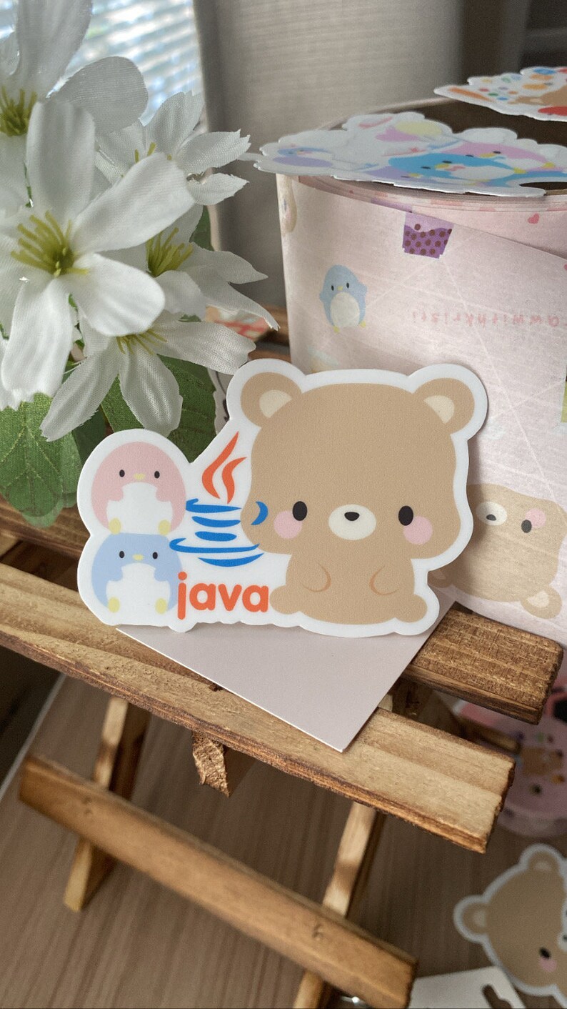 May include: A sticker featuring a cartoon bear and two penguins, with the word "java" in orange. The bear is light brown with pink cheeks. The penguins are pink and blue. The sticker is on a wooden surface.