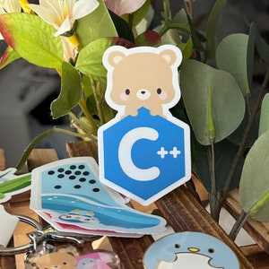 Könnte beinhalten: Eine Sammlung von Aufklebern und Schlüsselanhängern mit niedlichen Cartoon-Designs. Ein hellbrauner Bärenaufkleber thront über einem blauen Sechseck mit den Buchstaben "C++". Weitere Aufkleber zeigen einen Pinguin und ein Wellenmuster. Ein Schlüsselanhänger mit Bär- und Erdbeer-Design ist ebenfalls zu sehen.