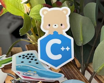 Puede incluir: Una colección de pegatinas y llaveros con diseños de dibujos animados. Una pegatina de oso marrón claro está colocada encima de un hexágono azul con las letras "C++". Otras pegatinas incluyen un pingüino y un diseño de ola. También se ve un llavero con un diseño de oso y fresa.
