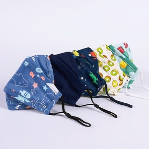 May include: Five colorful face masks with different patterns. The masks are made of fabric and have adjustable ear loops. The patterns include a blue mask with sea creatures, a navy blue mask with stars, a white mask with kiwi fruit, a green mask with a dinosaur, and a blue mask with a rocket ship.