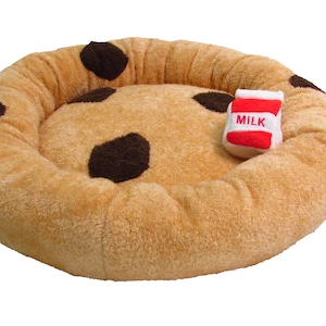 May include: A plush pet bed shaped like a giant chocolate chip cookie, with brown spots on a tan background. Includes a small, red and white milk carton toy. The word "MILK" is printed on the carton.